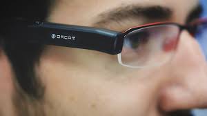 A profile view of a person using OrCam technology attached to their glasses. The OrCam and glasses are in focus and the person's face is blurred.