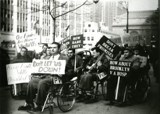 A group of people in wheelchairs holding signs on the streets of Manhattan. They are protesting for equal rights for disabled people. The photo is black and white.