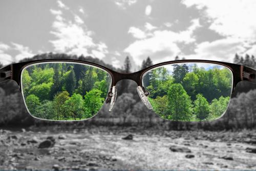A landscape that's out of focus and colorless with a pair of glasses in the middle - through the lenses everything is colorful and in focus