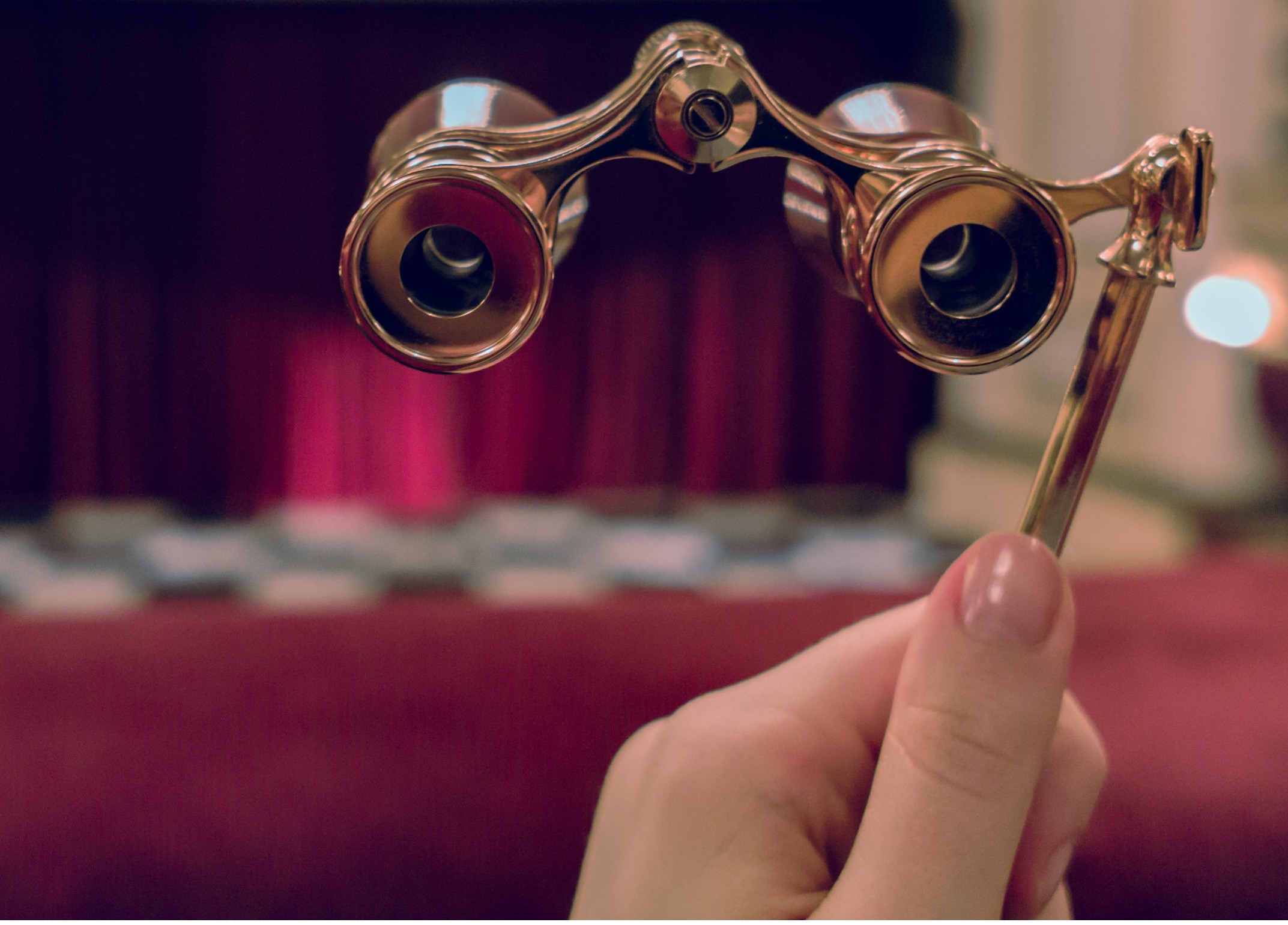 A picture of a person's left hand holding a pair of opera glasses in focus. Out of focus in the background is the stage and front rows of an opera house.