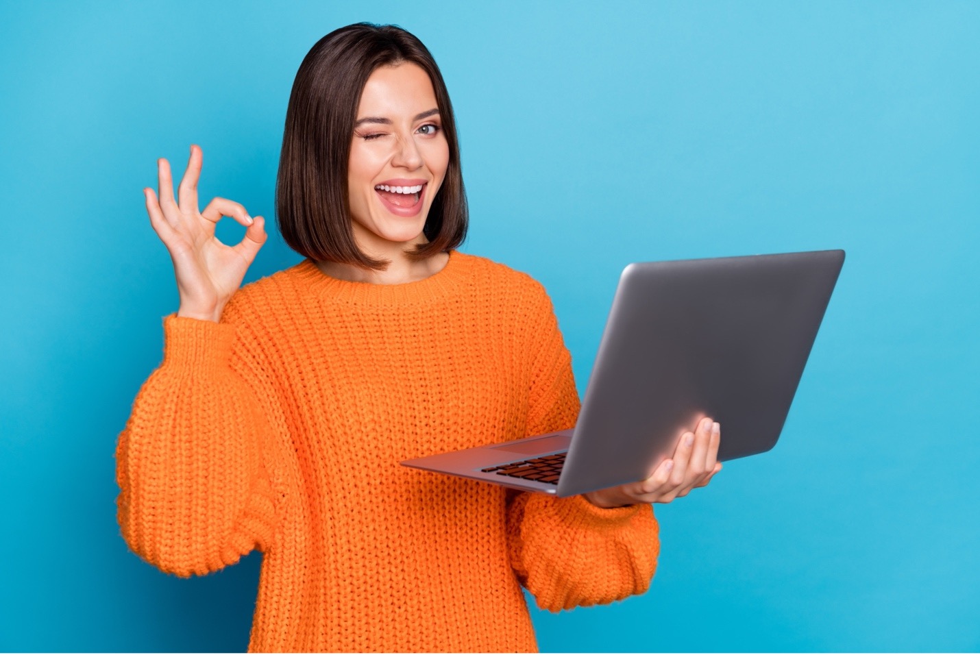 A woman holding a laptop in one arm, smiling, winking and holding up an "ok" sign with her other arm.