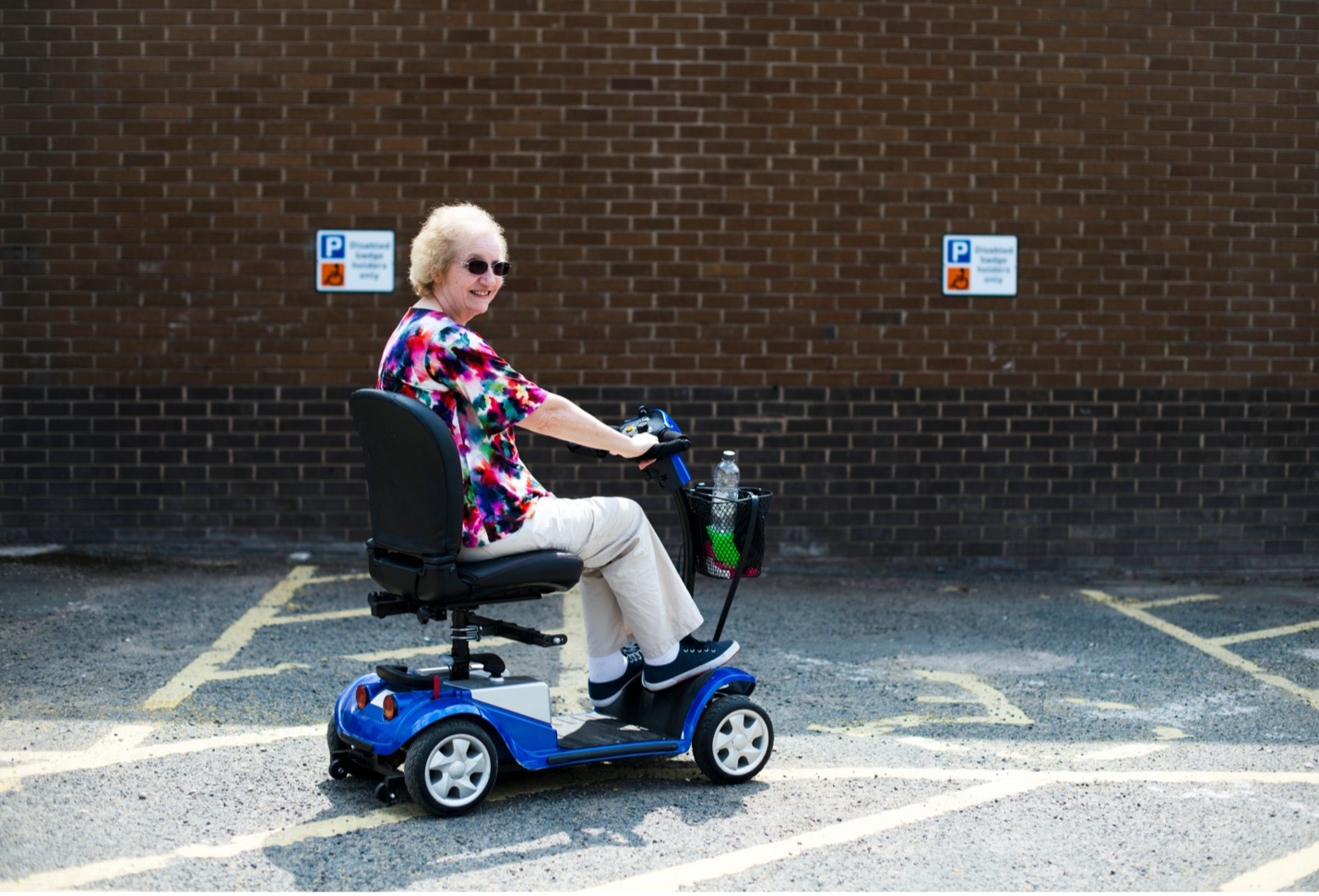 A smiling elderly woman on a motorized scooter in a parking lot on a sunny day