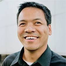 A picture of Jennison Asuncion - a light skinned man with black hair smiles at the camera. He wears a collared black shirt and is in front of a white background.